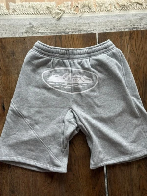 Grå sweatshorts med tryck - Säljer ett par grå sweatshorts med elastisk midja och ett vitt tryck framtill. Shortsen har en avslappnad passform och är tillverkade i mjuk bomullsmix. Bakficka och sidofickor för extra funktionalitet. Perfekta för chill dagar.