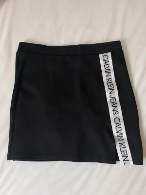 Svart kortkjol från Calvin Klein - Svart kortkjol från Calvin Klein med vit logotypremsa längs ena sidan och liten CK-detalj bak. 