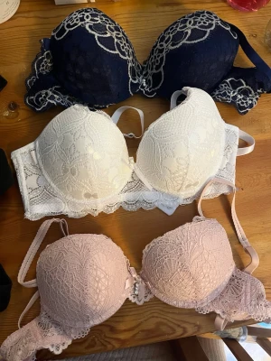 3-pack spetsbhar i olika färger - Säljer ett set med tre bhar i spets: en marinblå, en vit och en ljusrosa. Alla har vadderade kupor, justerbara axelband och dekorativa spetsdetaljer. Den ljusrosa har en liten rosett och pärlberlock framtill. Perfekt för dig som gillar romantisk och feminin stil. Säljer alla för 90kr men pris kan diskuteras 30kr/styck men säljer alla tillsammans det står storlek 70A men passar nog större eftersom de är ganska stora aldrig använda 