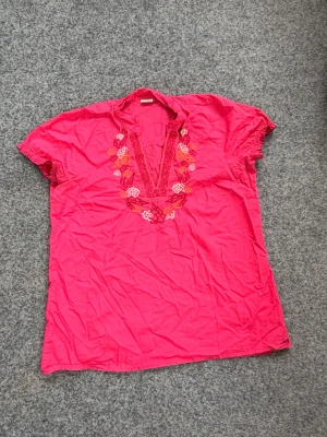 Rosa blus med broderi från Esprit - Säljer en rosa blus från Esprit med v-ringning och broderade blommor. Blusen har korta ärmar med små volangdetaljer och är gjord i ett lätt bomullstyg. 