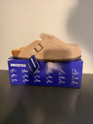 Beige Birkenstock Boston tofflor - Säljer ett par klassiska Birkenstock Boston tofflor i beige mocka med justerbart spänne och korksula. Perfekta för dig som vill ha både stil och komfort. Tidlös design som passar till det mesta och ger en avslappnad vibe.