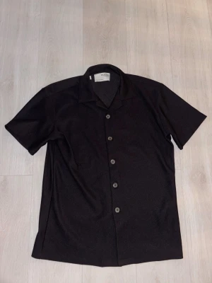 Svart skjorta från Selected Homme - Kortärmad svart skjorta från Selected Homme med klassisk krage och knappar framtill. Skjortan är tillverkad i ett ribbat material som ger en snygg struktur och avslappnad känsla. Perfekt till jeans eller shorts för en clean look.