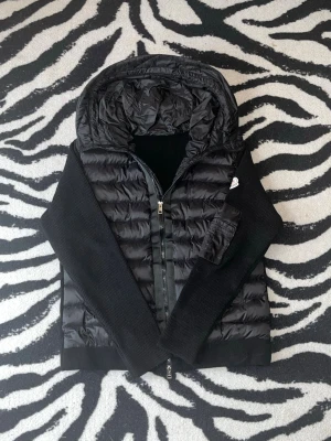 Moncler Cardigan - Snygg svart cardigan från Moncler med stickade ärmar och huva. Jackan har dragkedja framtill, ribbade muddar och en ficka med dragkedja på ärmen. Moncler-logga på vänster ärm. Perfekt för kalla vinterdagar eller till hösten. Pris kan diskuteras.
