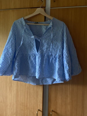 Blåvitrutig blus med knytning - Supersöt blus i blåvitt rutigt tyg med två knytband framtill och vidd i ärmarna. Blusen har en avslappnad passform och är tillverkad i ett lätt, krinklat material som känns luftigt. Perfekt för en trendig och avslappnad look.