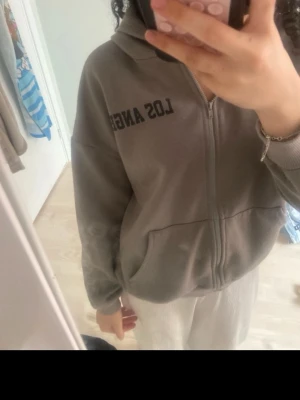 Grå hoodie med dragkedja och tryck - Säljer en grå hoodie med dragkedja och huva. På bröstet står det 'LOS ANGELES' i svart text. Hoodien har två stora fickor framtill och ribbade muddar vid ärmsluten. Perfekt för en chill och avslappnad stil.