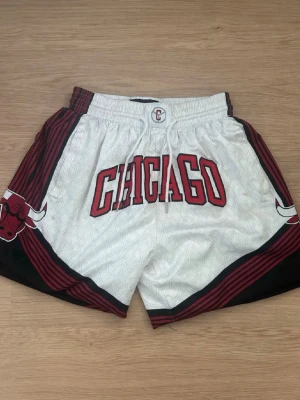 Chicago Bulls vita basketshorts - Snygga vita basketshorts med stort CHICAGO-tryck framtill och Chicago Bulls-logga på sidorna. Shortsen har röda och svarta detaljer, elastisk midja med snörning och fickor bak. Perfekta för dig som gillar streetwear och sportig stil.