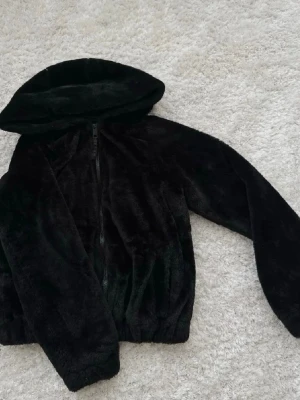 Svart teddyjacka med huva - Mysig svart päls jacka med dragkedja framtill och stor huva. Jackan har långa ärmar och resår i nederkant för en skön passform. Tillverkad i mjukt, fluffigt material som ger en trendig look och håller dig varm under kyliga dagar.