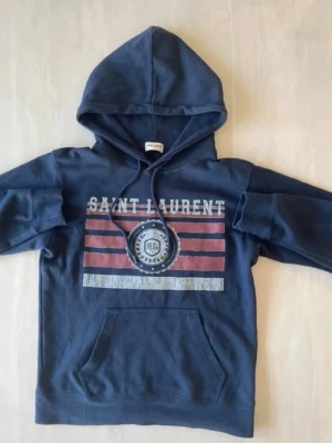 Saint Laurent hoodie - Snygg saint Laurent hoodie med huva och magficka. Framsidan har ett tryck med texten 'SAINT LAURENT' och grafiska ränder i rött och vitt. Perfekt för en avslappnad streetwear-look.
