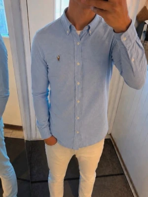 Ljusblå skjorta med knappar - Snygg ljusblå skjorta med button-down krage och vita knappar hela vägen. Skjortan har lång ärm och en liten broderad detalj på bröstet. Perfekt till jeans eller chinos för en clean och stilren look.