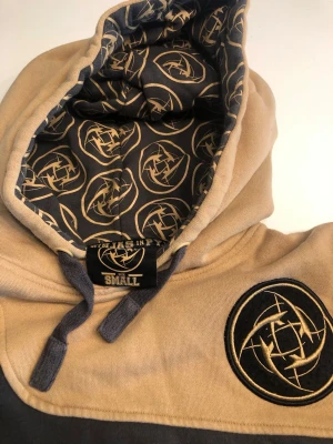 Ninjas in Pyjamas hoodie beige/grå - Säljer en hoodie från Ninjas in Pyjamas i beige och mörkgrå färg. Hoodien har ett stort tryckt logomärke på bröstet och mönstrat foder i huvan med lagets logga. Dragsko i huvan och ribbade muddar. Perfekt för dig som gillar gaming och streetwear.