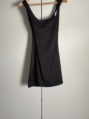 Svart kort satin-klänning - Stilren svart kort klänning i satin med breda axelband och figurnära passform. Perfekt för dig som gillar minimalistisk och elegant stil. Klänningen har en enkel design utan extra detaljer och ger en snygg siluett.