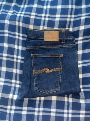 Nudie jeans  - Snygga mörkblå jeans från Nudie Jeans med klassisk femficksdesign och kontrastsömmar. Jeansen har en rak passform och är tillverkade i denim med tydliga detaljer på bakfickan. Perfekta för en stilren look. OBS uppsydda för att motsvara L30! . JAG SKICKAR EJ FLER BILDER NÄR JAG BÄR JEANSEN! för bättre inblick i passformen rekommenderar jag att googla på modellnamnet. För storleksguide kolla måtten noggrant då jeansen alltid kan vara uppsydda heller krympta och jag ej tar emot returer :)