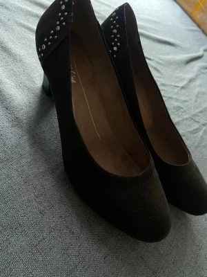 Svarta pumps med nitar - Säljer ett par svarta pumps med rund tå och mellanhög klack. Skorna har dekorativa silverfärgade nitar på hälen och är gjorda i ett mockaliknande material. Perfekta för dig som vill ha en stilren men ändå edgy look.
