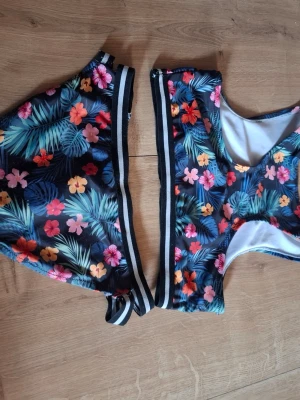 Blommig bikini med svarta detaljer - Snygg bikini med tropiskt blommönster i rosa, gul, orange och blå toner. Både överdel och underdel har svarta och vita ränder längs kanterna. Perfekt för sommaren och strandhäng!