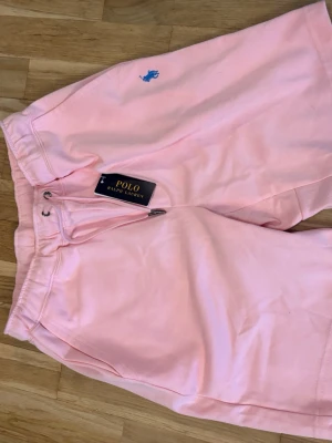 Rosa shorts från Polo Ralph Lauren - Snygga ljusrosa shorts från Polo Ralph Lauren med blå broderad logga på ena benet. Shortsen har snörning i midjan och är tillverkade i mjuk bomull, perfekta för sommardagar. Klassisk och avslappnad passform.