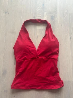 Röd halterneck linnetopp - Snygg röd halterneck linnetopp med djup v-ringning och åtsittande passform. Toppen har breda band som knyts bakom nacken och är tillverkad i ett mjukt bomullsliknande material. Perfekt för en trendig och avslappnad look.