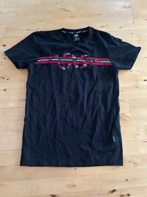 Svart t-shirt med orm och ränder - Svart t-shirt från Black Squad med färgglada ränder i rött och grönt över bröstet och en stor orm broderad ovanpå. Klassisk rund hals och korta ärmar. Tillverkad i mjuk bomull som känns skön mot huden.