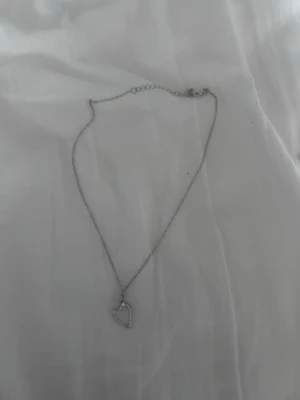 Silverfärgat halsband med hjärthänge - Ett stilrent halsband i silverfärg med en tunn kedja och ett öppet hjärta som hänge. Perfekt för dig som gillar minimalistiska smycken och vill ha något enkelt men ändå snyggt. Halsbandet är i metall och passar till många olika stilar.