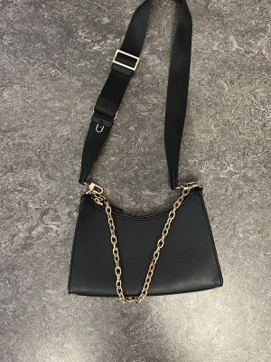 Svart handväska med kedjedetalj - Stilren svart handväska i slätt konstläder med guldfärgad kedja och justerbar axelrem i textil. Väskan har en minimalistisk design med dragkedja upptill och passar perfekt för dig som gillar trendiga accessoarer.