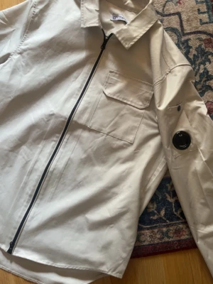 Beige overshirt från C.P. Company - Stilren beige overshirt från C.P. Company med dragkedja framtill, klassisk krage och bröstficka. Ärmen har den ikoniska svarta linsen med logga. Perfekt lager för en trendig streetwear-look.
