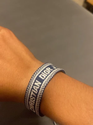 Blått broderat armband Christian Dior - Snyggt blått och vitt armband från Christian Dior med broderad logga. Armbandet är gjort i textil och har justerbar knytning med tofsar i ändarna. Perfekt accessoar för att lyfta din stil med en lyxig touch.