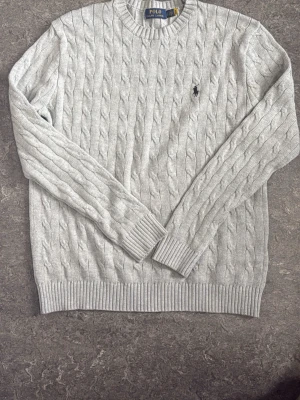 Grå kabelstickad tröja Polo Ralph Lauren - Snygg grå kabelstickad tröja från Polo Ralph Lauren med klassisk rund halsringning och svart broderad logga på bröstet. Tröjan har långa ärmar och ribbade muddar vid ärmslut och nederkant. Perfekt för lager på lager och stilren vardagslook.