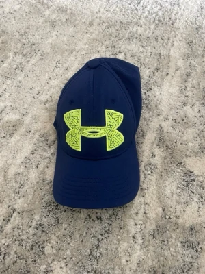 Blå keps från Under Armour - Snygg blå keps från Under Armour med ett stort neongult logotyptryck framtill. Skärmen är böjd och undersidan är också neongul. Kepsen har ventilerande hål och justerbar passform. Perfekt för en sportig och avslappnad stil.