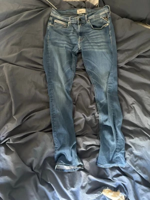Blå jeans från Replay - Snygga blå jeans från Replay med klassisk femficksmodell och ljusa slitningar framtill. Jeansen har normal midja och raka ben. Tillverkade i ett mjukt denimtyg som ger en bekväm känsla. Perfekta för dig som gillar en avslappnad men stilren look.