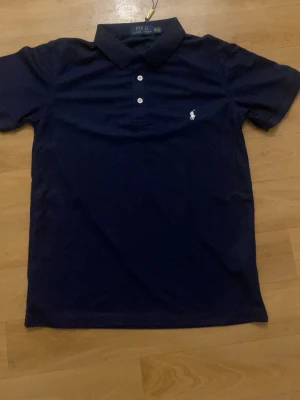 Marinblå pikétröja från Ralph Lauren - Klassisk marinblå pikétröja från Ralph Lauren med vit broderad logga på bröstet och två vita knappar framtill. Tröjan har korta ärmar och mjukt bomullsmaterial som känns skönt mot huden. Perfekt för en stilren och avslappnad look.