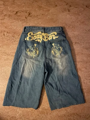 Blå denimshorts med tryck bak - Säljer ett par blå denimshorts med bred passform och gult tryck på bakfickorna och baktill i midjan. Shortsen har klassisk femficksdesign och är tillverkade i denim. Perfekta för dig som gillar streetwear/y2k och vill sticka ut lite extra.