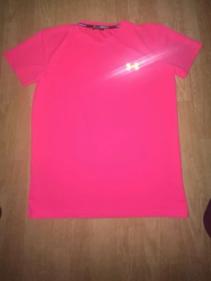 Rosa t-shirt från Under Armour - Snygg rosa t-shirt från Under Armour med klassisk logga på bröstet. Perfekt för träning eller chill, tillverkad i ett lätt och mjukt material som andas. Kortärmad modell med normal passform.