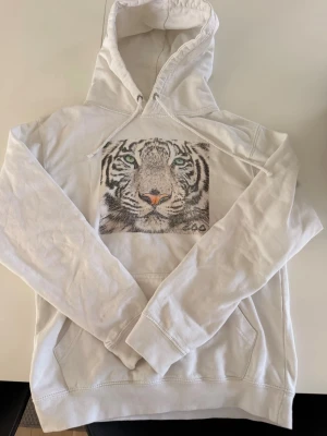 Vit hoodie med tigertryck - Säljer en vit hoodie med stor huva och dragsko. På bröstet finns ett detaljerat tryck av ett tigeransikte med gröna ögon och orange nos. Hoodien har magficka och ribbade muddar. Perfekt för dig som gillar djurmotiv och vill sticka ut.