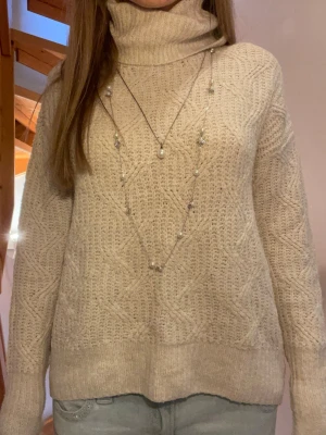 Beige stickad polotröja - Mysig beige stickad polotröja med snyggt mönster och hög krage. Tröjan har långa ärmar och ribbade muddar vid ärmslut och nederkant. 