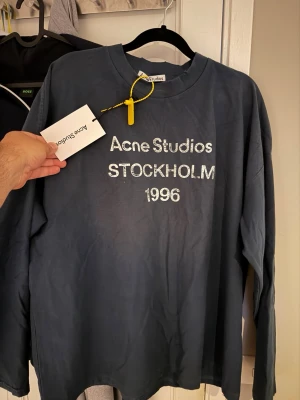 Blå långärmad t-shirt Acne Studios - Snygg blå långärmad t-shirt från Acne Studios med tryck 'Acne Studios STOCKHOLM 1996' på bröstet. Tillverkad i mjuk bomull och har en avslappnad passform. Perfekt för en stilren och trendig look.