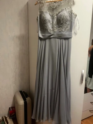 Ljusgrå/silvrig långklänning med spetsdetaljer - Elegant ljusgrå långklänning med vackra spetsdetaljer och broderier upptill. Klänningen har ärmlös design och transparent mesh vid axlarna. Kjolen faller mjukt och ger en svepande siluett. Perfekt för speciella tillfällen. Använd en gång! Även en scarf tillkommer med den. 