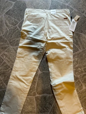 Beige chinos från Jack & Jones - Snygga beige chinos från Jack & Jones i modellen JJIMARCO JJBOWIE SA. Byxorna har raka ben, två bakfickor med knapp och är tillverkade i ett mjukt bomullsmaterial. Perfekta för en clean och stilren look.