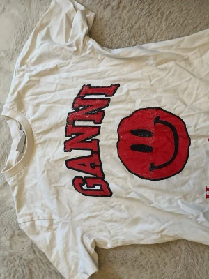 Vit Ganni t-shirt med smileytryck - Vit t-shirt från Ganni med stort rött smileytryck och svart text på framsidan. förlåt om den är lite skrynklig men jag ska strycka den!!❤️