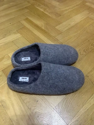 Gråa tofflor från ASOS DESIGN - Mysiga gråa tofflor från ASOS DESIGN i mjukt filtat material. Klassisk slip-in modell med rundad tå och fluffigt foder på insidan för extra komfort. Perfekta för att hålla fötterna varma hemma.