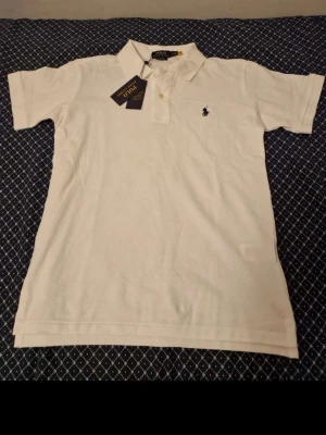 Vit pikétröja från Polo Ralph Lauren - Klassisk vit pikétröja från Polo Ralph Lauren med broderad logga på bröstet. Tröjan har korta ärmar, krage och knappar framtill. Tillverkad i bomull för en skön och luftig känsla. Perfekt till jeans eller shorts för en clean look.