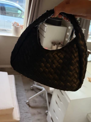 Svart flätad axelväska - Snygg svart handväska i flätad design med rundad form och kort axelrem. Väskan har dragkedja upptill och är tillverkad i ett material som liknar konstläder. Perfekt för dig som gillar trendiga accessoarer och vill ha en unik detalj till din outfit.