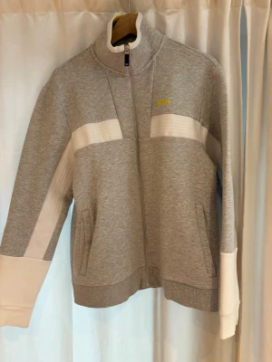 Grå zip hoodie från BOSS - Snygg grå hoodie från BOSS med dragkedja framtill och vita detaljer på ärmar och bröst. Tröjan har hög krage, två fickor och ribbade muddar. Tillverkad i mjukt sweatshirtmaterial som är perfekt för en avslappnad stil. Och ett gult Boss märke på bröstet