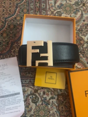 Svart Fendi bälte med guldfärgat spänne - Snyggt svart bälte från Fendi med ett stort, guldfärgat F-spänne. Bältet är tillverkat i skinn och levereras i originalask med tillhörande kvitto och förpackning. Perfekt accessoar för att lyfta din outfit.