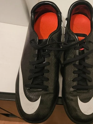 Svarta Nike fotbollsskor med vit swoosh - Snygga svarta fotbollsskor från Nike med stor vit swoosh på sidan och röda innersulor. Skorna har klassisk snörning och är tillverkade i ett slitstarkt syntetmaterial som ger bra bollkontroll. Perfekta för dig som vill sticka ut på planen. 