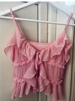 Rosa volang topp med knyt - Supersöt rosa linnetopp med tunna axelband och volangdetaljer. Toppen har knyt framtill och är gjord i ett lätt, plisserat tyg som ger en luftig känsla. Perfekt för varma dagar och ger en romantisk vibe till din outfit.
