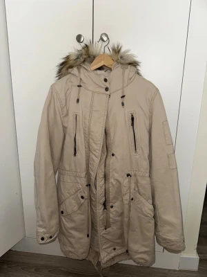 Beige parkas med fuskpäls - Stilren beige parkas med stor huva och avtagbar fuskpäls. Jackan har flera dragkedjeförsedda fickor, justerbar midja och knappar vid ärmslut. Perfekt för kalla dagar och ger en snygg streetwear-look.