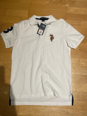 Vit pikétröja från U.S. Polo Assn. - Klassisk vit pikétröja från U.S. Polo Assn. med korta ärmar och broderad logga på bröstet. Tröjan har krage, knappar vid halsen och en mörk detalj på ärmen. Perfekt för en clean och sportig stil.