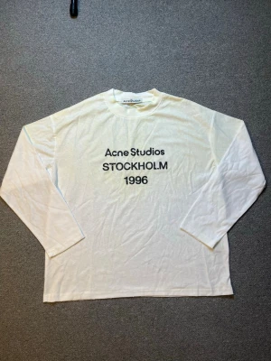 Vit långärmad topp Acne Studios - Vit långärmad topp från Acne Studios med svart text 'Acne Studios STOCKHOLM 1996' på bröstet. Klassisk rak passform och rund halsringning. Perfekt för dig som gillar minimalistisk och stilren design.
