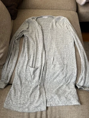 Ljusgrå lång kofta från Name It - Mysig ljusgrå lång kofta från Name It med två framfickor och öppen front. Tillverkad i mjukt, tunt material som passar perfekt för lager på lager. Enkel och stilren design som funkar till det mesta.