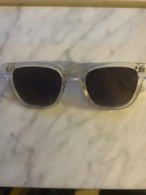 Transparenta solglasögon med svarta glas - Snygga solglasögon med transparent båge och mörka, svarta glas. Klassisk fyrkantig form som ger en modern och stilren look. Perfekta för att lyfta din outfit under soliga dagar. Köpta från XXL men inte säker på märket.