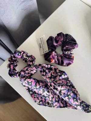 Blommiga scrunchies - Två olika modeller av blommiga scrunchies i lila och rosa. Ena är knappt använd och den andra (i sammet) har prislapp kvar på.